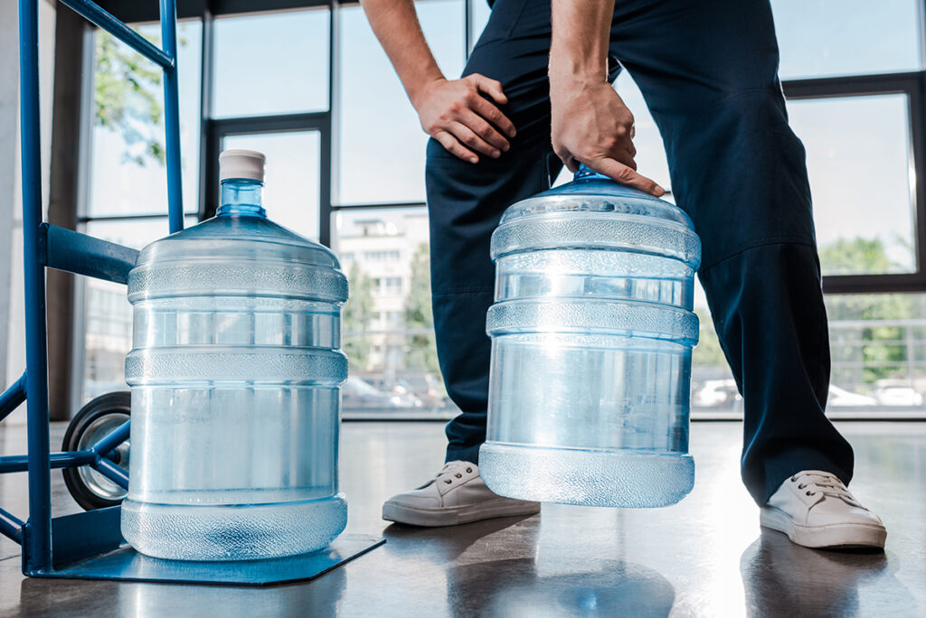 Close up of heavy water cooler jugs taking up floor space. A man is lifting one of the jugs off of the floor