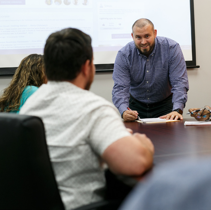 Man in a checkered shirt leans over a conference table, presenting to colleagues as slides project on a screen behind him.