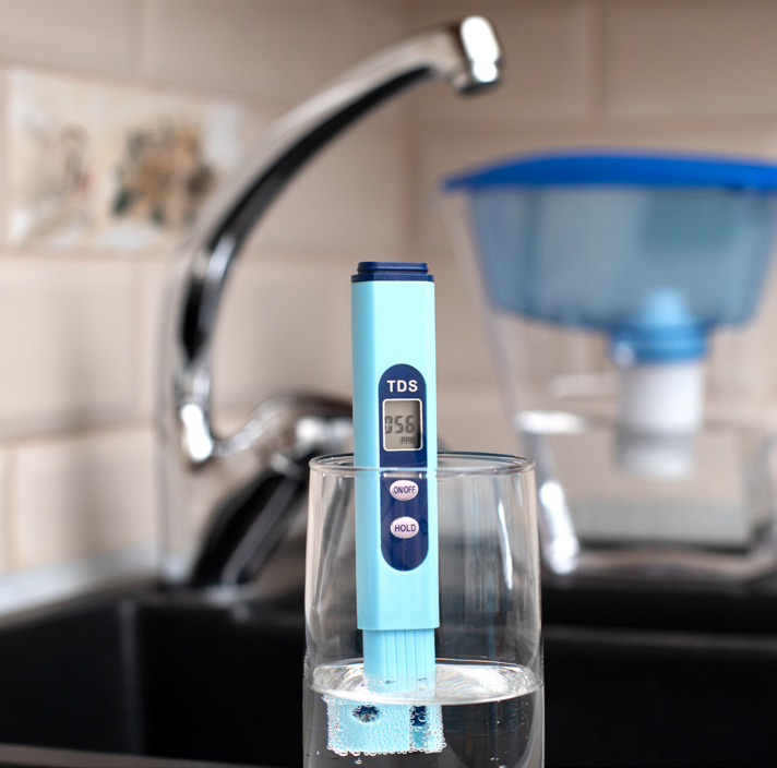 Blue TDS water tester immersed in a glass of water on a kitchen counter, showing a reading of 058 ppm. This describes water quality measurement rather than the device’s appearance.