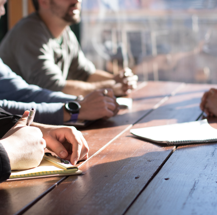 People seated at a wooden table taking notes with pens and notebooks during a meeting.
