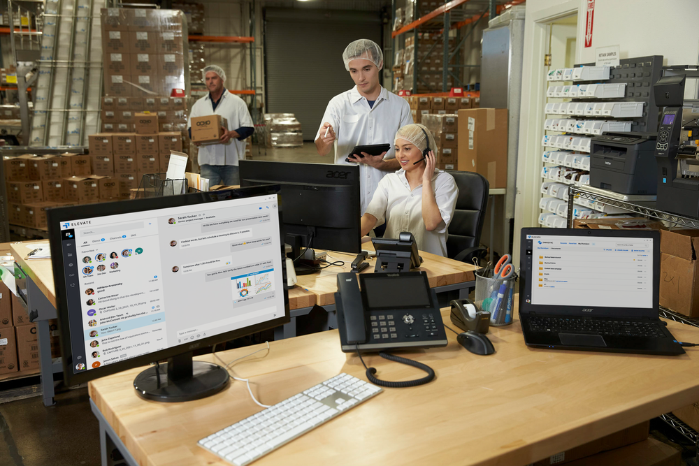 desks in a factory with computers and office phone. People working in background
